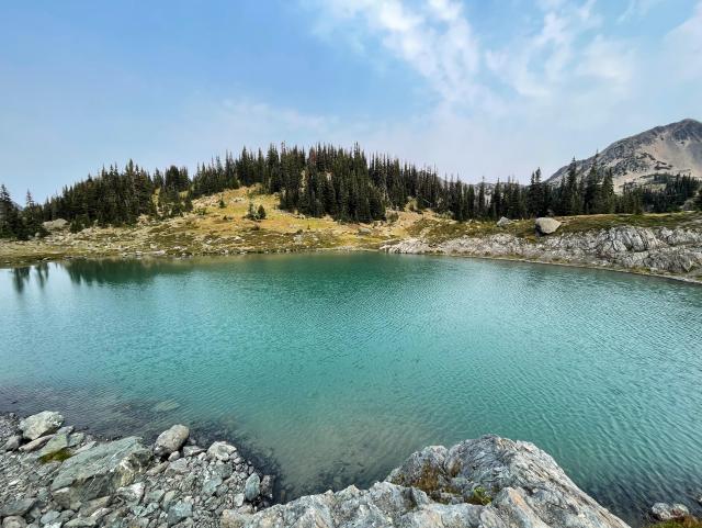 A small turquoise, perfectly clear, body of water in a rocky basin with wind-driven ripples.  Across the water is a hill with conifers and grey mountains.   The Imperial Tarn in Olympic National Park.