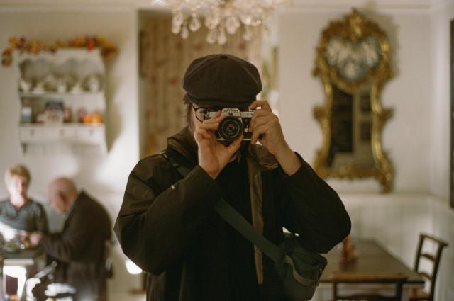 A man wearing a flat cap, a black wind jacket and an over the shoulder pouch, holding a Pentax KX camera with both hands in front of his face, reflected in a mirror. In the background, a glass chandelier, antique shelves and wooden tables in the interior of Brady's Tearoom in Grasmere. An elderly couple is sitting at one of the tables.