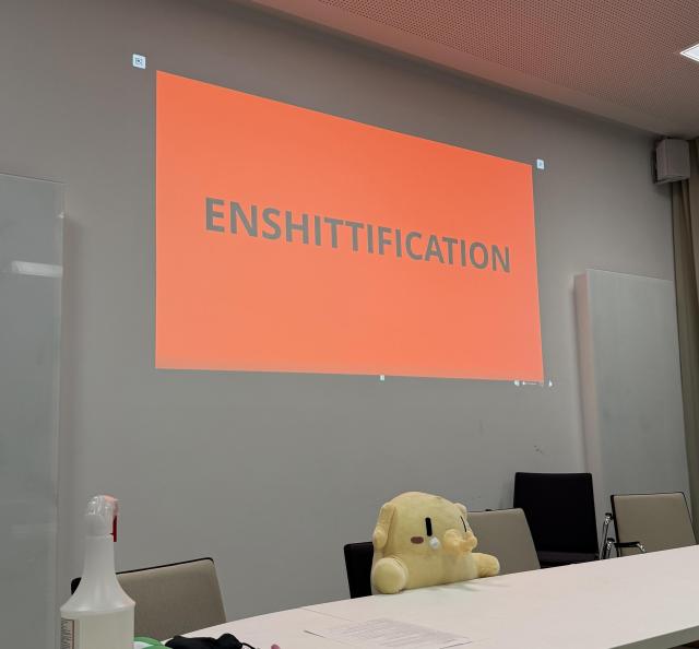 Man sieht eine Projektionsfläche in Rot mit dem Wort "Enshittification". Davor sitzt der Plushtodon und es sieht aus, als ob er den Talk gibt.