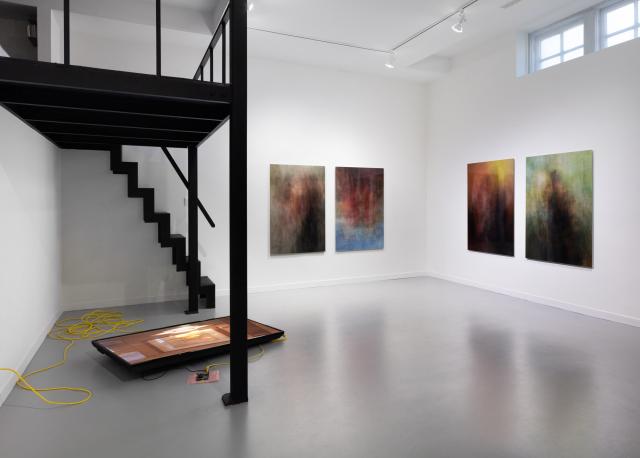 A full exhibition view. In the foreground is a tv lying flat on the floor surrounded by yellow cables. On the walls are four large photographic prints.