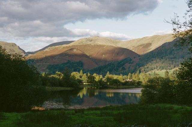 A calm shaded lake reflecting green trees illuminated by golden sunlight. In the distance, fells covered in auburn colored ferns.