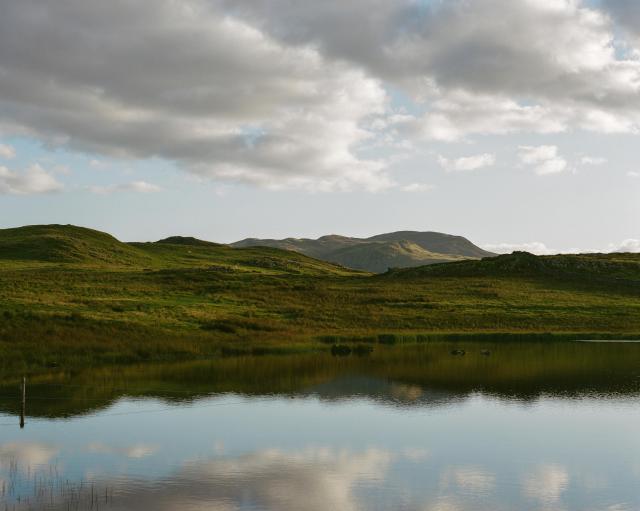 Lightly rippled water reflecting the grass and fells on the opposite side.