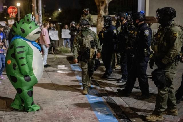 Person in an inflatable frog costume faces down a line of armed security forces in front of the ICE facility in Portland, Oregon.