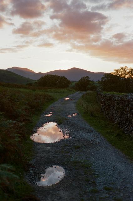 A gravel path next to a dry-stone wall under a pink sky. There are puddles reflecting the sky. In the distance, sun setting behind the fells.