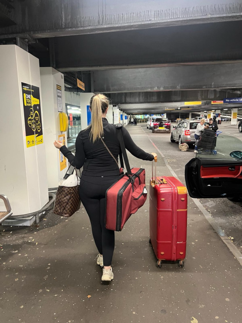 Iona stands in the parking bay at airport drop off with red nord piano bag over shoulder and red suitcase and hair in pony 