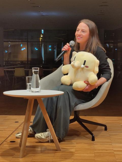 Melanie Bartos sitzt auf einer Bühne in Innsbruck mit einem Mikrofon in der einen Hand. In der anderen Hand das Maskottchen von Mastodon, wie es hier an der Universität Innsbruck heisst Federike.