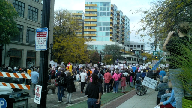 Looking West: A large crowd of people marching down a city street during a protest, holding signs, with a tall apartment building in the background and a "No Parking Snow Route" sign visible.