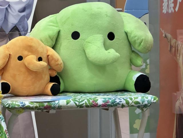 Two plush elephant toys sit on a floral-patterned surface. One is orange and small, while the other is larger and green. Both have simple features with round eyes and a curved trunk.