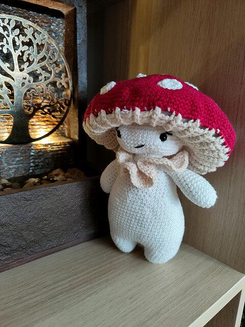 Crocheted plush toy resembling a whimsical mushroom character. The body is white with short, rounded arms and legs, and the head is topped with a large red mushroom cap featuring evenly spaced white spots. The underside of the cap is textured in light beige to mimic gills, matching a ruffled collar around the neck. The face is minimalistic, with two small black stitched eyes and a subtle smiling mouth. The toy leans against a wooden surface beside a decorative metallic item adorned with small stones.