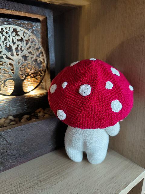 Handmade crocheted plush toy styled as a mushroom character, viewed from behind. The toy has a white stem-like body with short legs and a large red mushroom cap adorned with evenly spaced white spots. The underside of the cap is not visible, but the rounded shape and proportions suggest a soft, stuffed structure. The toy is positioned in a corner beside a decorative light display featuring a metallic tree design and small stones at the base. Warm lighting from the display casts a gentle glow, highlighting the texture and craftsmanship of the plush toy.