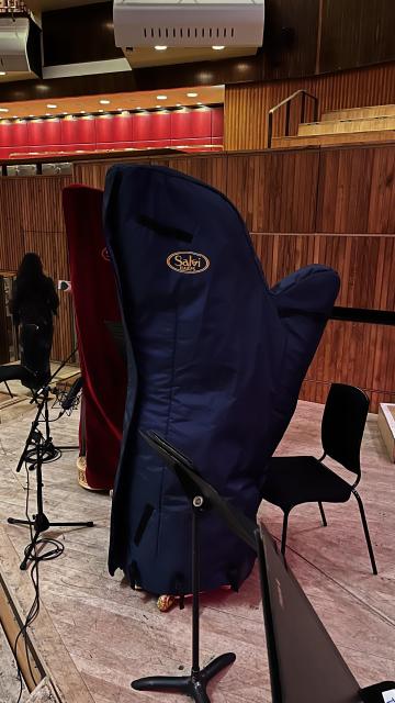 The image shows two harp covers in a concert setting, one in blue and one in red, positioned on a wooden stage. There's a music stand and a black chair visible, with a person partially in the background.