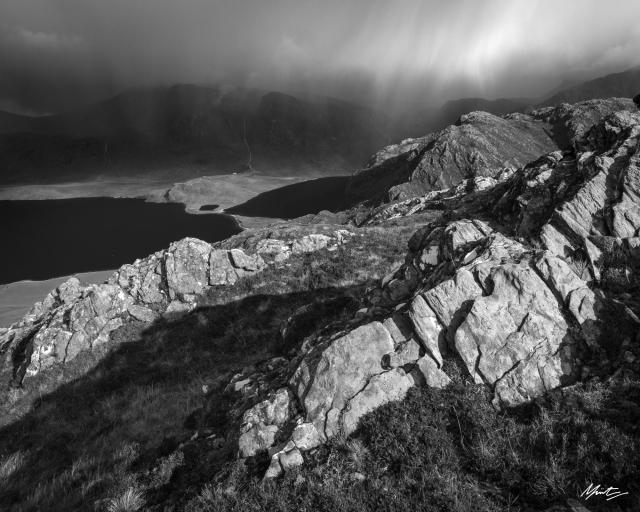 a black and white mountain photograph from the scottish highlands. gneiss rocks catching sunlight sweep diagonally in the foreground while a rainstorm approaches over mountains and distant lochs in the back ground. overall, a dramatic moody scene