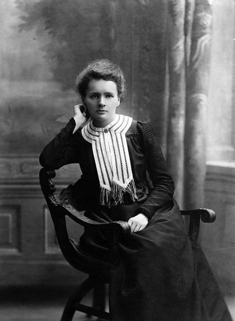 Black-and-white portrait of Maria Skłodowska-Curie, 1903. She is seated in a chair, wearing a dark dress with a light, patterned collar, resting her head on one hand and looking directly at the camera. public domain
