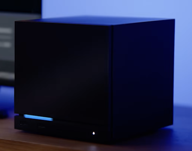 Small black cubic desktop computer or external drive with a thin blue LED strip and a small round power indicator, sitting on a wooden surface against a blue-lit background.