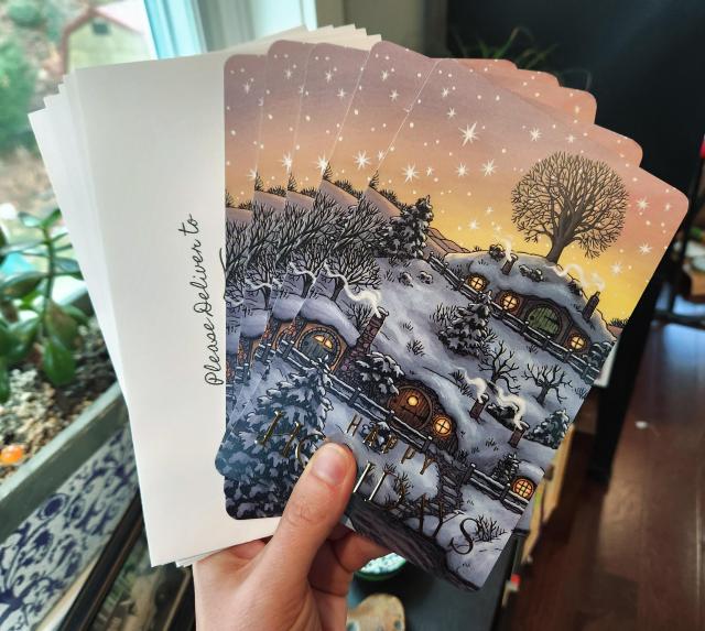 Photograph of the greeting cards held in a hand, along with envelopes. The greeting card features an illustration of Hobbiton covered in snow, with inviting warmly lit windows, smoke coming from chimneys, and snow covered and bare branch trees.