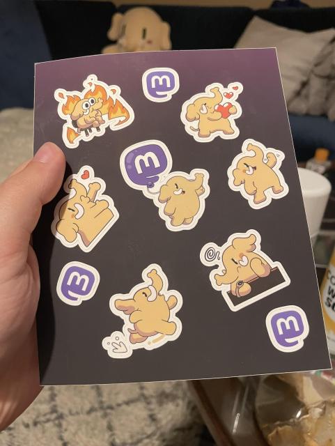 A sheet of stickers featuring the Mastodon mascot in various situations, as well as 3 Mastodon logos, held in my hand. 