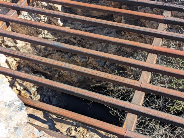 A large rectangular mine shaft in a rocky desert region, covered with a rusty metal grate 