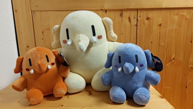 All three Mastodon plushies are sitting next to each other. The original yellow one which is roughly twice as big as the other blue and orange ones in the middle.