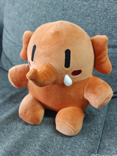 Das Mastodon Stofftier Maskottchen - in der Farbvariationen "Activity Peach" (dunkles orange) - sitzt auf einer grauen Couch