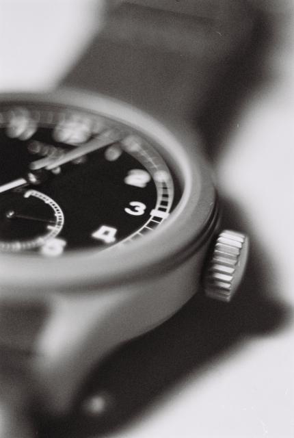 A grainy and blurry, black-and-white macro photograph of a very non-dressy wristwatch. The watch is a reissue of a British dirty dozen field watch from World War II, with their characteristic black face and white numerals, a railroad minutes track and a seconds sub-dial, a fairly bulky bezel and a plain but generously knurled crown. It even has the broad arrow symbol on the dial, blurred and hiding behind the minutes hand.