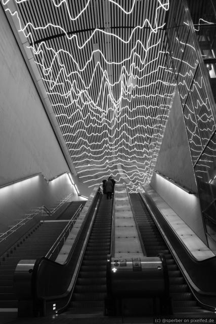 Black-and-white image taken from the bottom of an escalator. There is an LED installation on the ceiling with white mountain shaped lines.