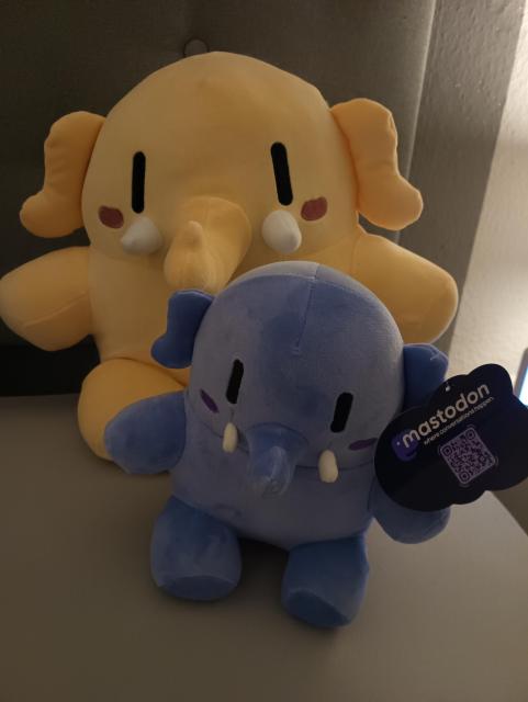 Ein kleines hellblaues Plüschtier in Form eines Mastodon sitzt vor dem größeren und hellgelben Ebenbild.