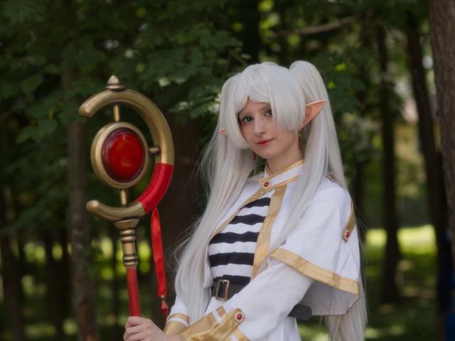 The image captures a young woman standing amidst a forested area. She is dressed in an elaborate costume that includes white hair, pointed ears suggesting elfin traits, and makeup around her eyes enhancing their appearance. The attire consists of a striped shirt with horizontal black stripes. Over this, she wears a long-sleeved jacket adorned with red buttons and golden trimmings.

In her hand is a staff that has a striking design: it's predominantly brass-colored with an eye-like motif in bright red embedded within a circular frame towards its top section. The woman holds the staff upright by its handle, which appears to be made of wood, contrasting against the metallic sheen above and below. Her gaze meets the camera, giving her character a sense of engagement.

The backdrop is lush greenery indicative of trees with dense foliage. This setting provides a natural contrast that highlights the fantastical elements embodied in the woman's costume.