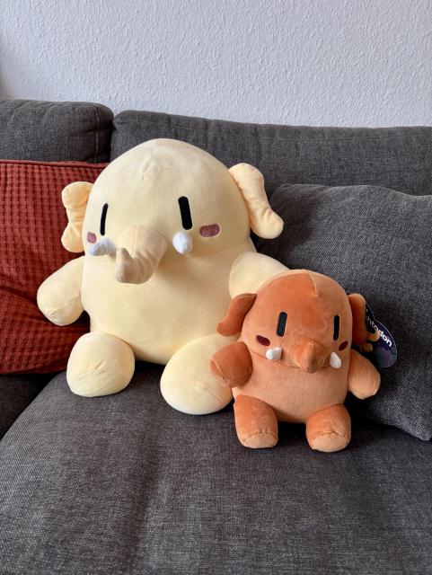 Two adorable plush elephant toys sit on a gray sofa. The larger plush is yellow with small black eyes and rosy cheeks, while the smaller one is orange with similar features. A red textured pillow is visible in the background.