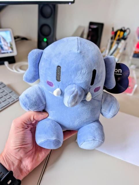A close-up of a hand holding a round, light blue plush elephant toy. The elephant has black oval eyes, small white tusks, and purple blush marks on its cheeks. A dark blue tag is attached to its ear. The background features a slightly blurred office desk with a computer monitor, keyboard, speaker, and various stationery items.