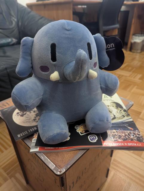 Mini Mastodon plushie Blue sitting on a small wooden stool in the living room. There's the most recent eclipsed magazine sticking out on the side, so it's more specifically sitting on that.