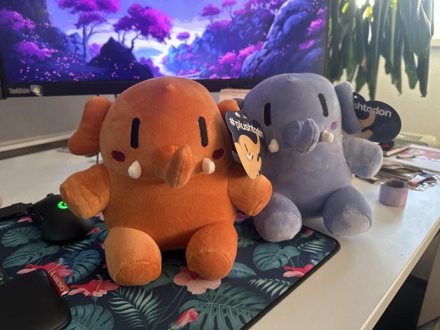 Two plush mastodon toys sit on a desk, one orange and one purple, both featuring tags that read "#plushtodon." A computer monitor with a colorful landscape background is visible behind them, along with a gaming mouse and a floral-patterned mousepad.