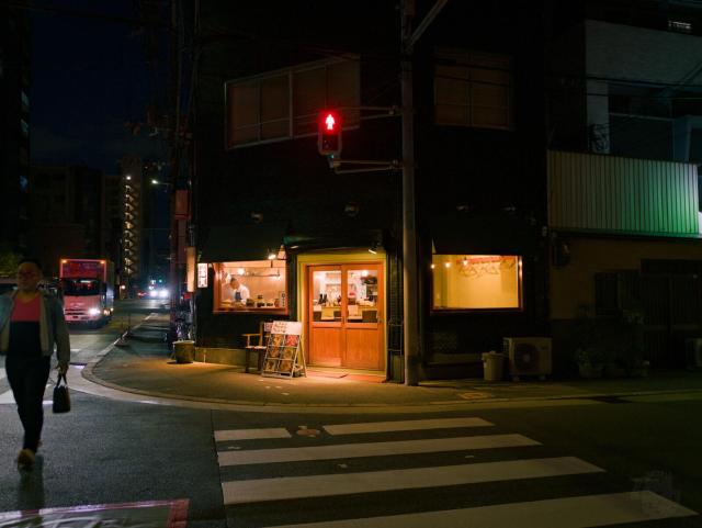 Nighttime urban street intersection. A building with a wooden door and illuminated windows sits at the corner. A pedestrian crosswalk with white stripes spans the road in the foreground. A person walks on the left side, wearing a pink top and dark pants, carrying a bag. A red traffic light is mounted on a pole near the building. In the background, a red truck is visible on the road, with distant buildings and streetlights under a dark sky. The sidewalk features a chair, a signboard, and a trash can.