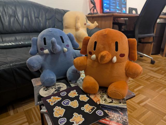 Blue and Peach Mini Mastodons sitting next to each other in a living room, looking at the camera. Sticker sheet in front of them. Yellow Adult Mastodon is sitting on a couch in the background, looking to the right