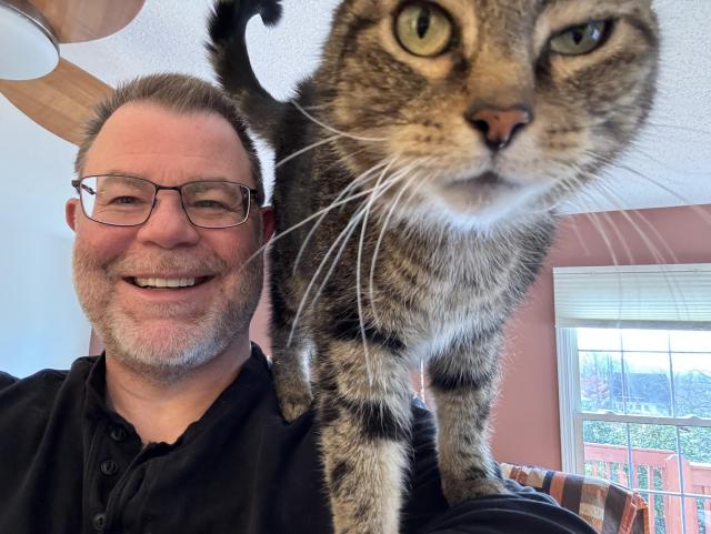 A middle-aged white guy with lots of grey in his beard and glasses, with a tabby cat on his shoulder putting its face right in the camera. 