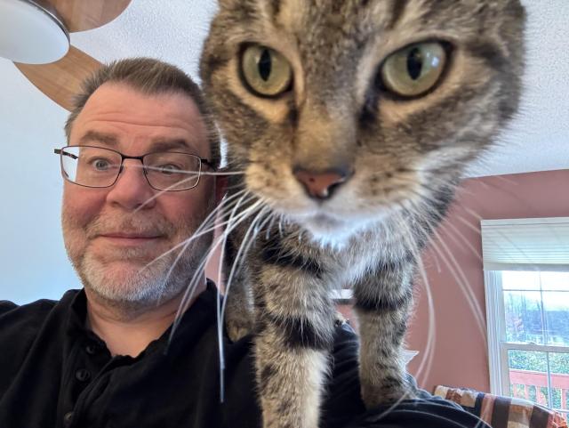 A middle-aged white guy with lots of grey in his beard and glasses, with a tabby cat on his shoulder putting its face right in the camera. 