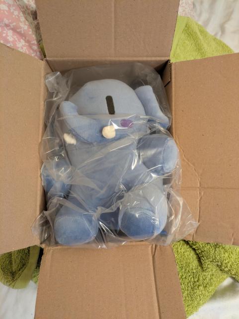 A small blue Mastodon plush still in the clear plastic bag, in a cardboard box