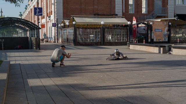 The image depicts a man crouched down on the sidewalk, holding his arm out towards an object in front of him, making a photo on his smartphone. The object appears to be a statue or sculpture, and it is situated between two buildings with large windows. A few people can be seen walking by in the background, adding life to the scene.