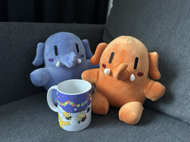Blue and orange Mastodon plushies, with a Mastodon mug, decorated with a winter scene, in front of it