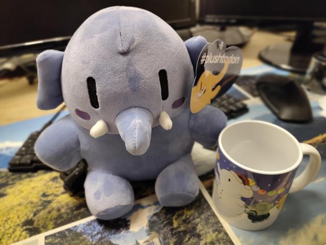 Ein blaues Plüsch Mastodon sitzt auf einem Schreibtisch, rechts daneben eine Mastodon Tasse mit Winter Design.