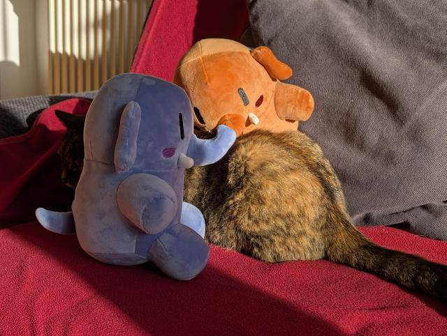 Same as photo 1, but the cat has turned her back away from the viewer, while the plushtodons are facing the cats' body looking very busy with their hands