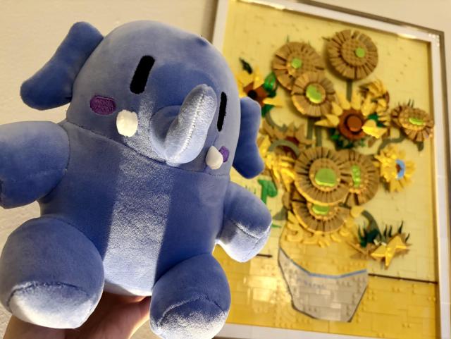 in focus is a dark blue Mastodon plushie, or stuffed animal, that i'm holding up. light is shooting up from below the frame, casting interesting shadows on the plushie. in the background is a bright yellow Lego recreation of a famous Van Gogh painting Sunflowers