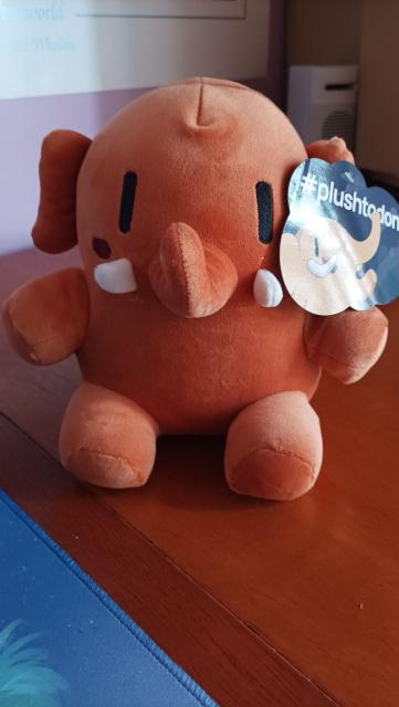 Photography of an orange mini Mastodon plushie on a desk. A plushtodon tag is attached to his left ear.