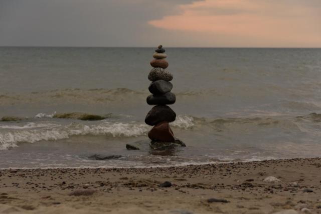 The image captures a serene scene at what appears to be dusk, with the sun setting in the background. A series of rocks are stacked meticulously on top of each other along the edge of a beach where calm waves lap against the shore. The sky above is painted with hues that suggest it's either dawn or sunset, adding warmth and tranquility to the scene. In the distance, one can see another body of water stretching towards the horizon under an expansive sky, suggesting this might be part of a larger coastal area.