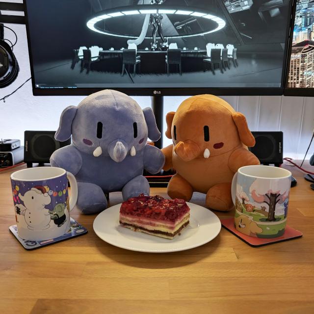 Two hungry looking Plushtodon plushies in blue and orange colors sit in front of a plate with a raspberry cake and two coffee mugs on each side.