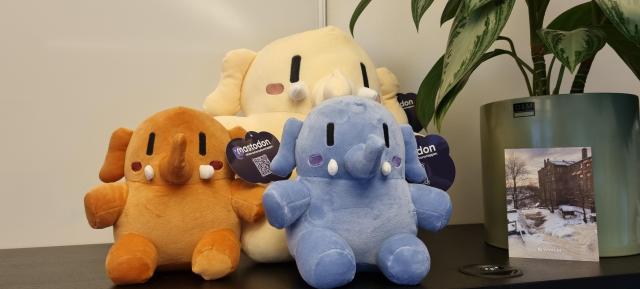 A photo of a trio of Plushtodons (Mastodon mascot plushies) sat on top of a black cabinet. Beside them is a potted plant and a postcard photo of the river next to the Vivaldi offices in winter.