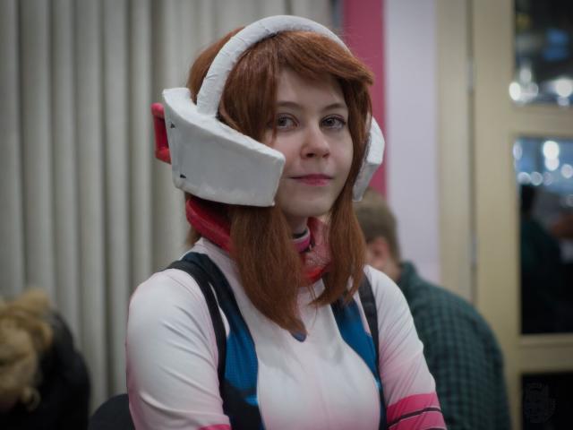 The image captures a moment of quiet contemplation. A young woman with vibrant brown-red hair stands out against the soft, blurred backdrop of what appears to be an indoor setting. She's wearing a white and pink hoodie adorned with blue sleeves, adding a pop of color to her outfit. Overhead, she is also wearing what appears to be a futuristic helmet with big blocks around ears, painted in bold red and white hues. The helmet has an open visor, revealing her focused gaze as if contemplating something significant. Her stance suggests she's standing still, absorbed by whatever lies before her.

The woman's face is turned towards the camera, giving us a clear view of her unique look. In the background, there are two other people partially visible behind her.