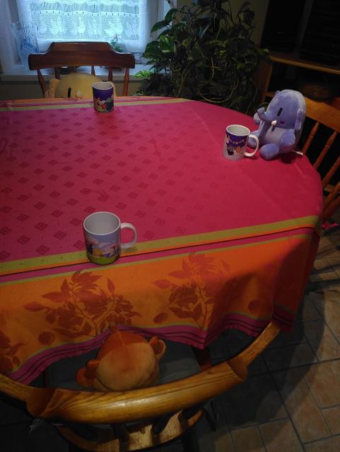 Photo mettant en scène les trois peluches Mastodon autour d'une table avec des chaises et les trois mugs Mastodon :
- le petit orange est assis sur une chaise avec la tasse Printemps, mais son regard ne dépasse pas le niveau de la table.
- le petit bleu est assis sur la table devant sa chaise avec la tasse Hiver, car il est malpoli.
- le grand jaune est assis sur une chaise avec la tasse Été, on voit la partie supérieure de sa tête péniblement dépasser du niveau de la table.
