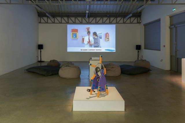 View of an exhibition space with a wall and a projection of the Energy Academy video with bean bags and a projector in front. In front of them there is a very colorful (yellow, blue, purple) perpetual motion machine.
