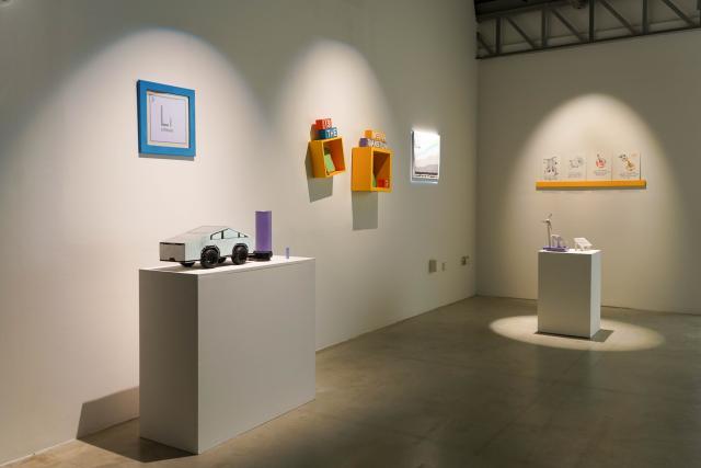 View of an exhibition space with white walls, there is a plinth with a DIY cybertruck and an illustration of the Lithium sign from the periodic table on the wall. Behind there are two bright yellow shelves with wooden blocks that spell our "Is the mission transition?" Behind them is another plinth with small objects and illustrations on the wall.