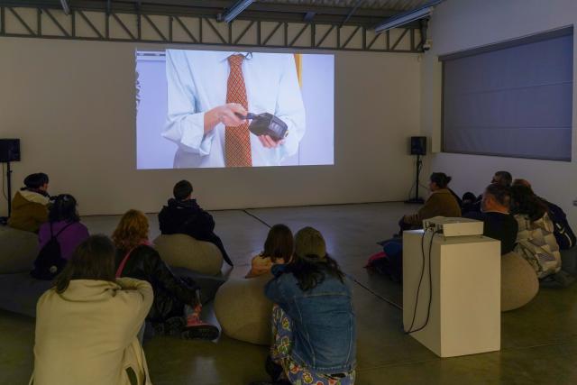 Same room as the first image with the same video projected on the wall, this time a dozen people are seated in front of the projection, watching the video with their backs towards the camera.